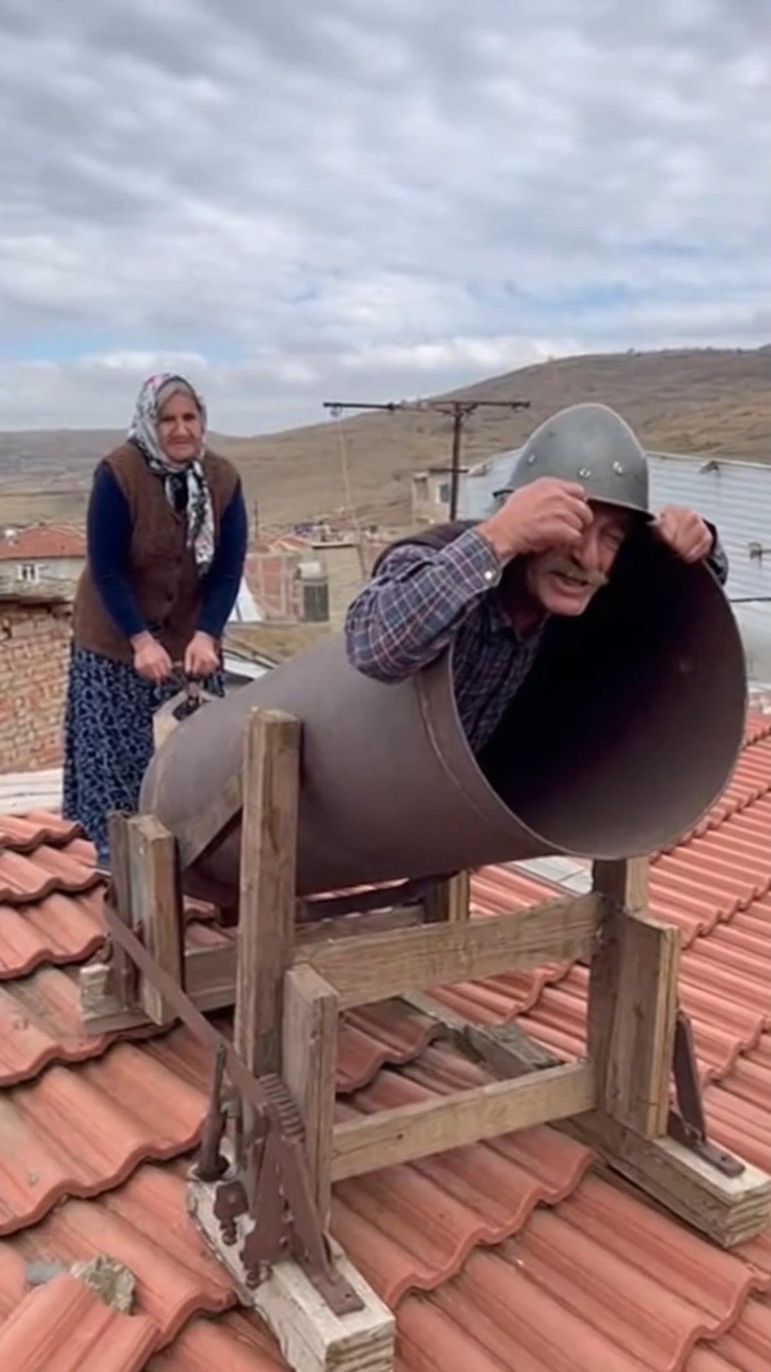 על גג בית בכפר, זוג נשוי מבוגר מכין תותח מאולתר. הגבר הזקן,...